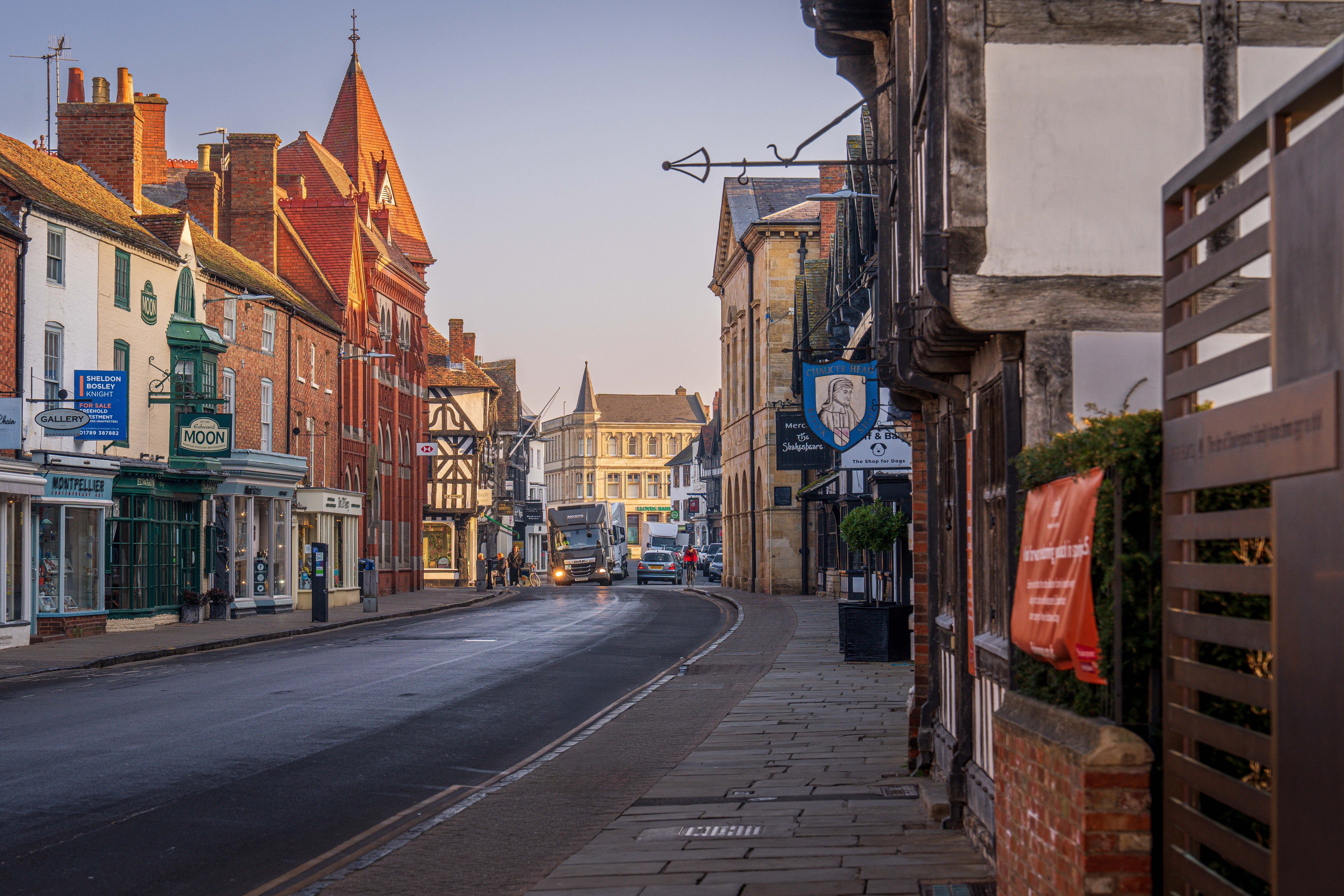 Half-timbered and red-brick buildings along a Stratford-upon-Avon street at dawn, with a Shakespeare pub sign