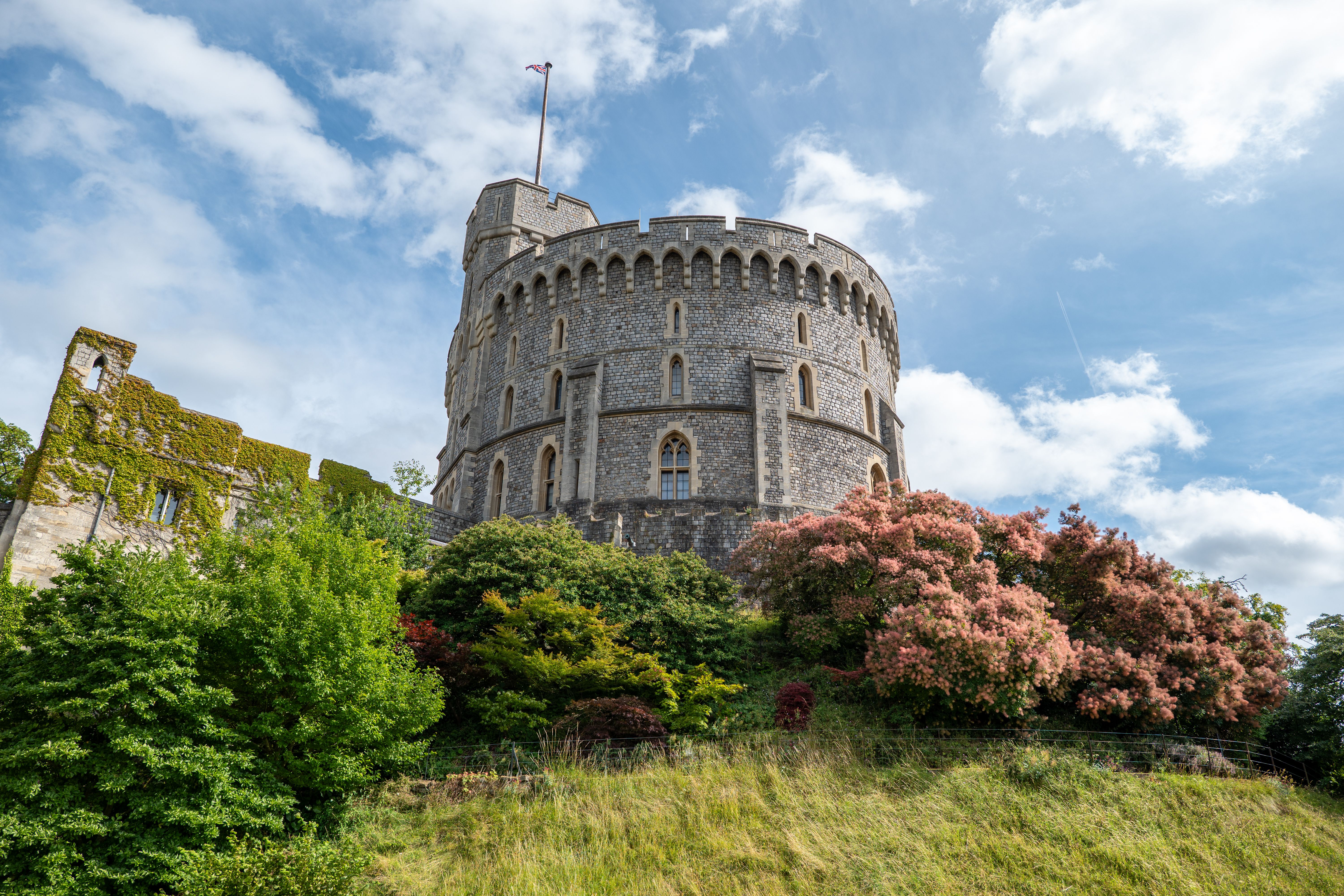 The Round Tower of Windsor Castle rising above green shrubs and a grassy slope under a blue sky