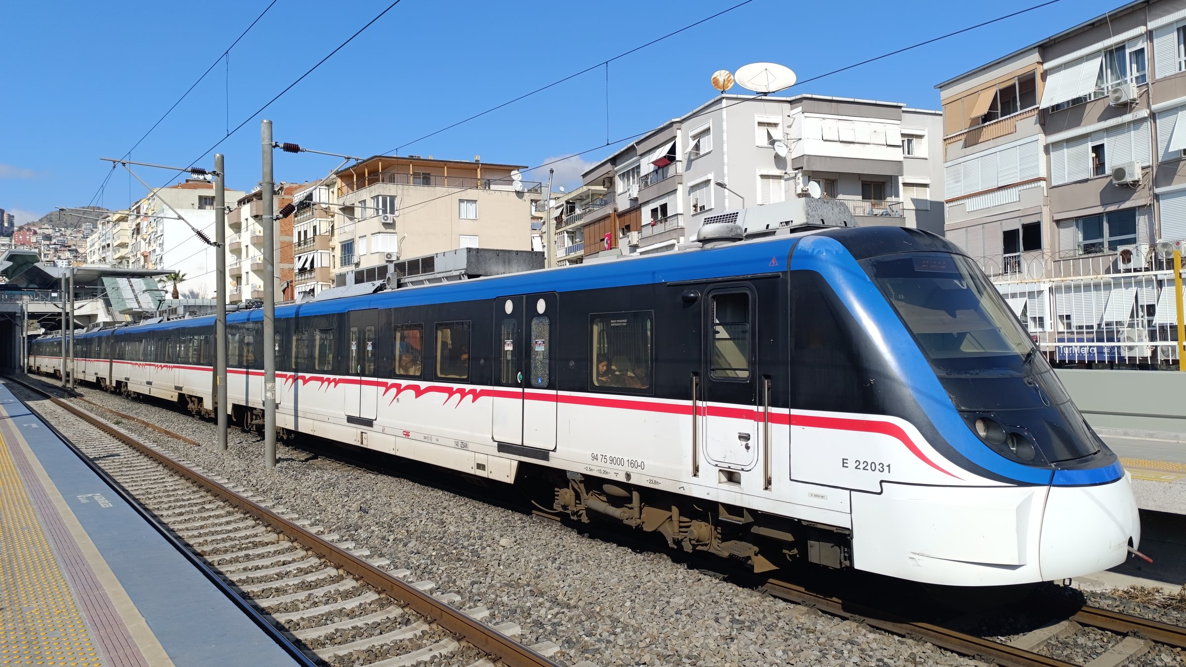 Un treno pendolare İZBAN blu, nero, bianco e rosso, unità E 22031, fermo a un binario di İzmir con palazzi e una collina alle spalle.