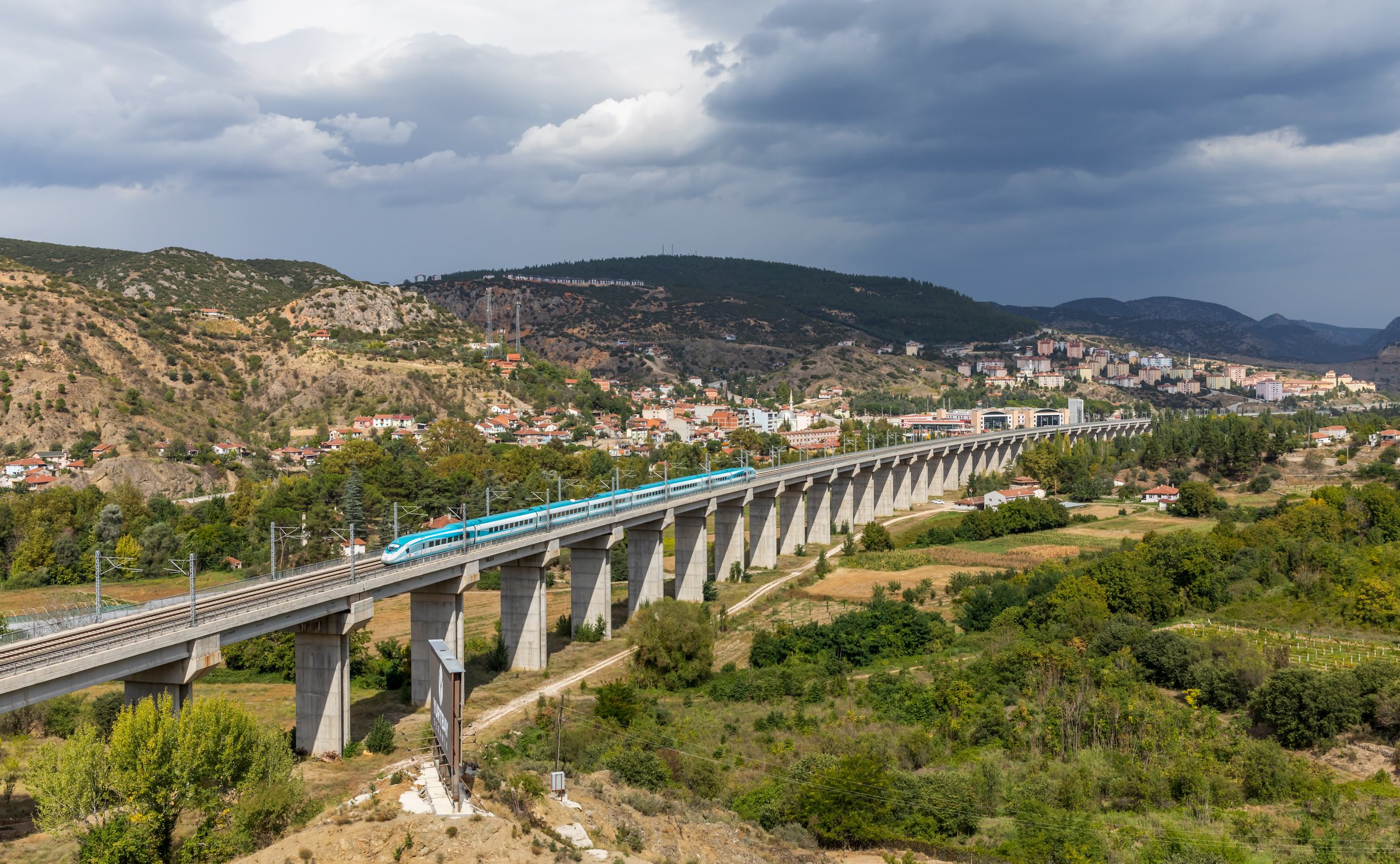 A white and turquoise TCDD Taşımacılık HT80000 high speed train crossing the long concrete viaduct above the town of Bilecik in north west Turkey.